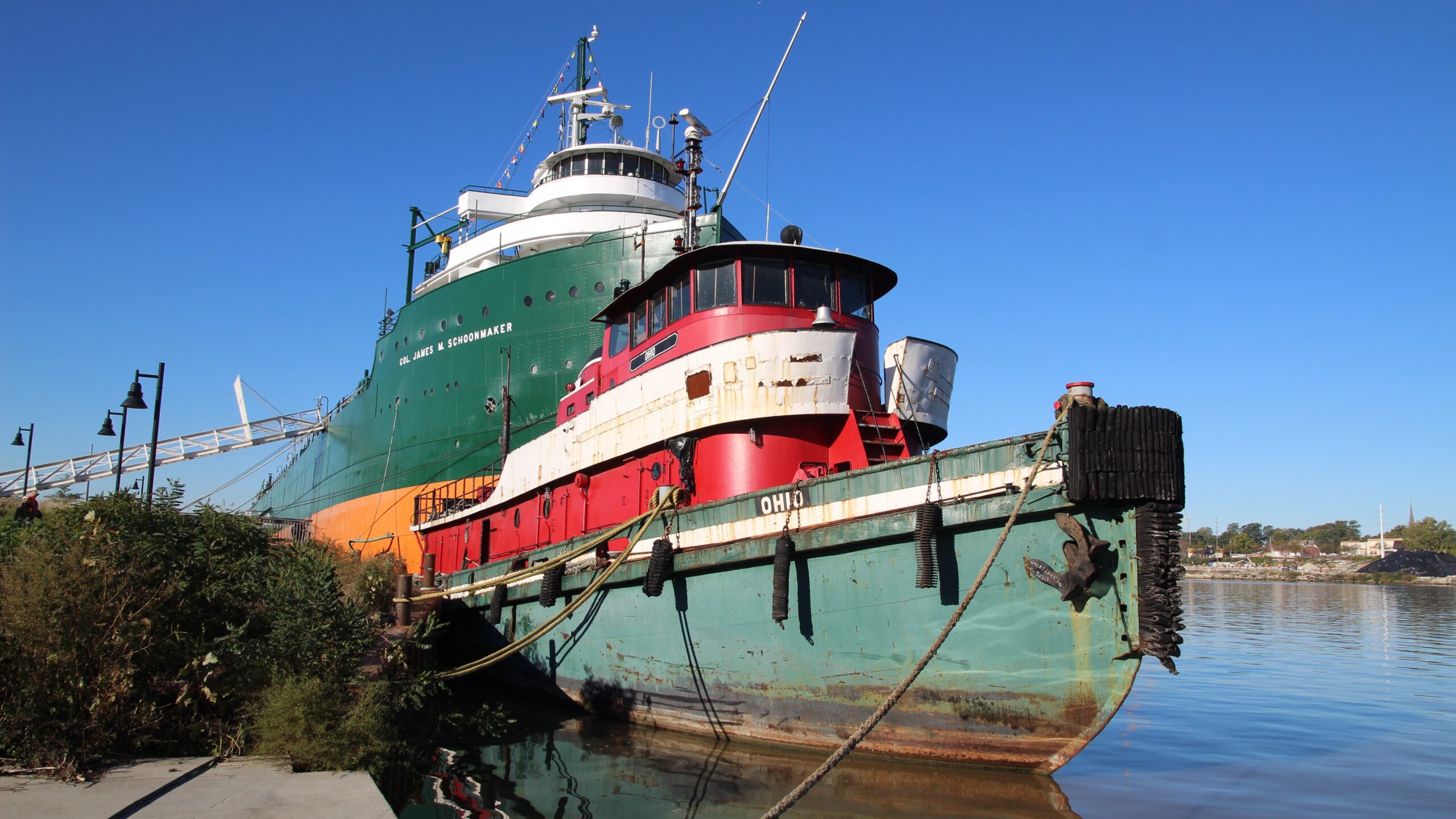 The red-and-green tugboat Ohio moored beside the freighter Col. James M. Schoonmaker under a clear blue sky, with the Maumee River behind them.
