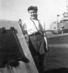 Bev is leaning against a monument. The lake and a boat are in the background. Bev is a young woman with her hair pulled back in a 1940s style with the top rolled back. She is wearing a white blouse tucked into loose wide-legged shorts.