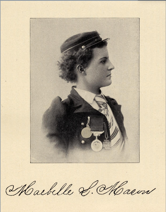 Maebelle is a teenage girl with short curly hair. She is wearing a dark cap and dark jacket over a white blouse and striped tie. There are three medals pinned to the front of her jacket.