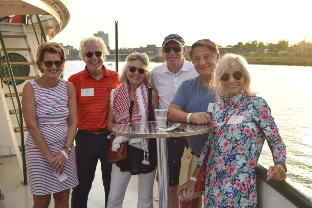A group of six adults in colorful clothing aboard the deck of a ship, drenched in warm sunset light.